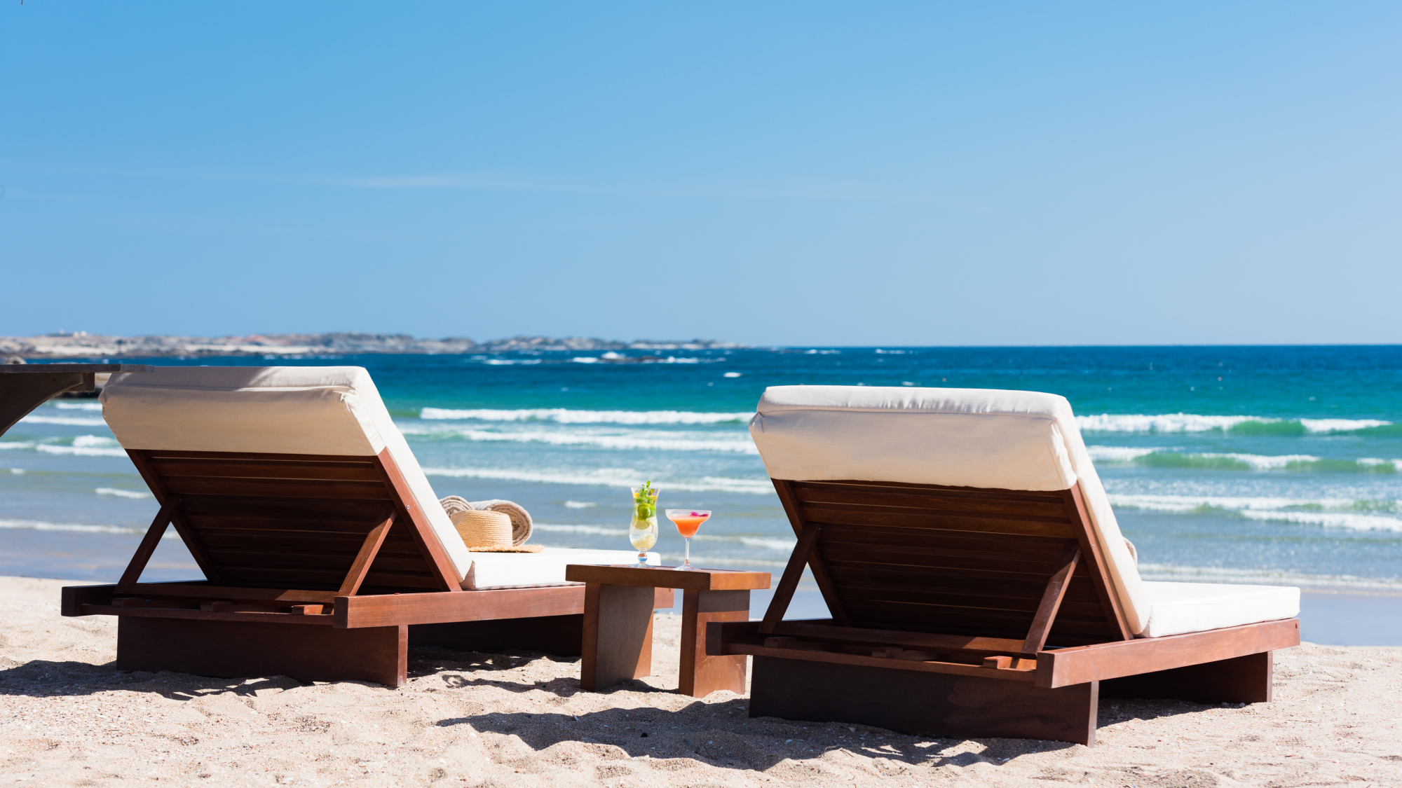 Two lounge chairs on the beach with two cocktails on a table between them.