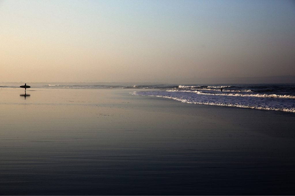 Person holding surfboard standing on the beach looking out at the ocean